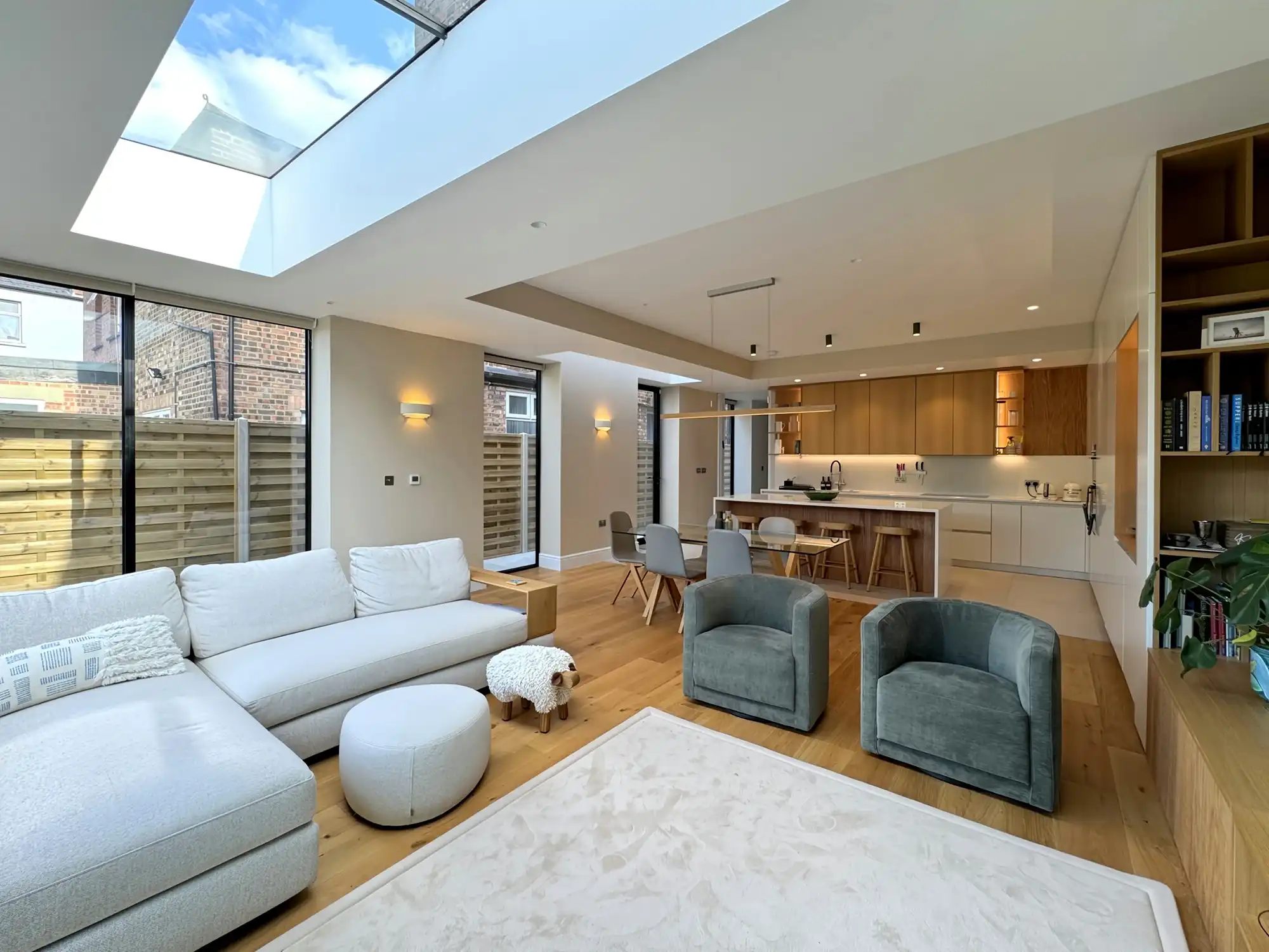 Modern open-concept living space featuring a gray sectional, round ottoman, two armchairs, and a sleek kitchen with large windows.